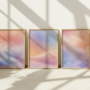 May include: Three framed art prints displaying abstract cloudscapes in pink, orange, and purple hues. The prints are in light-coloured wooden frames, leaning against a white wall. The artwork creates a feeling of peace and tranquility.