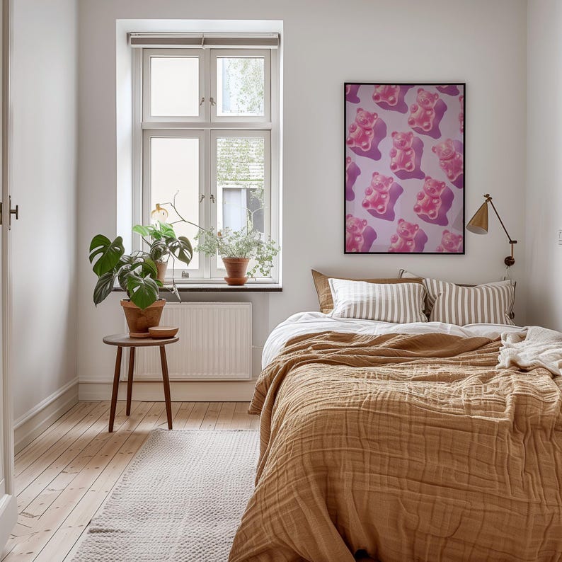 Pode incluir: Interior de quarto com uma cama coberta por um cobertor texturizado cor de mostarda e almofadas listradas. Um p&ocirc;ster emoldurado com ursinhos de goma rosa est&aacute; pendurado acima da cama. Uma pequena mesa de madeira suporta uma planta em vaso.