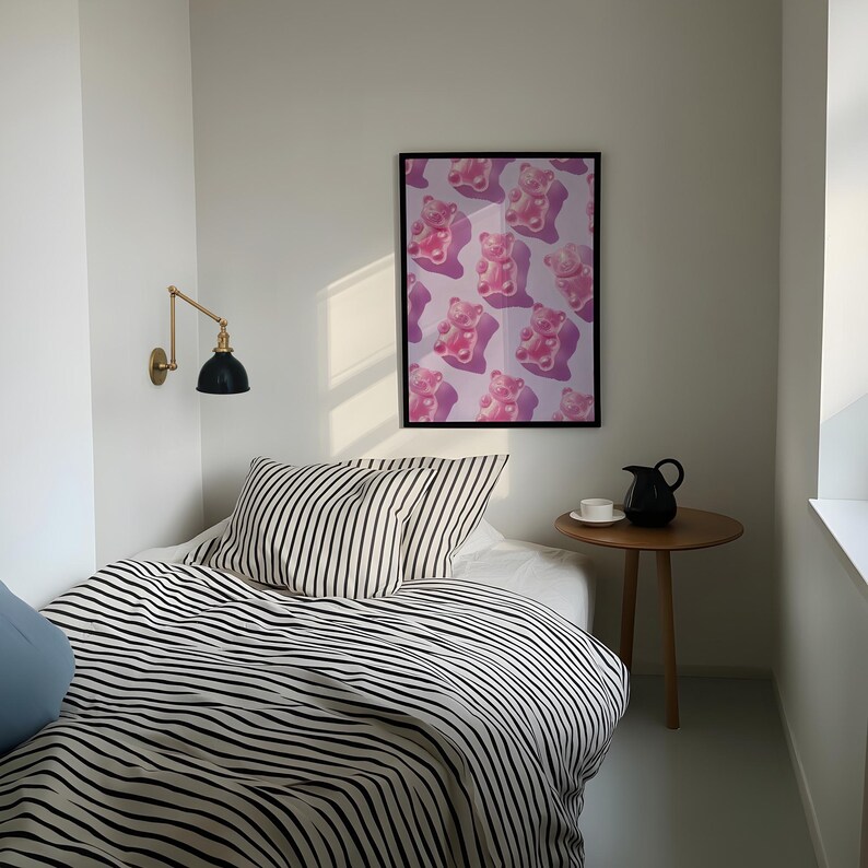 Pode incluir: Um quarto com uma cama com roupa de cama listrada em preto e branco e uma almofada azul clara. Uma impress&atilde;o emoldurada de ursinhos de goma rosa est&aacute; pendurada na parede. Uma pequena mesa lateral de madeira com um jarro preto e uma x&iacute;cara branca.