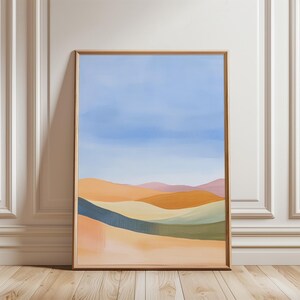 Könnte beinhalten: Abstrakter Landschaftskunst-Druck in einem Holzrahmen. Das Kunstwerk zeigt sanfte Hügel in Orange-, Gelb- und Grüntönen mit blauem Himmel. Das Gemälde wird vor einer weißen Wand und einem Holzboden ausgestellt.