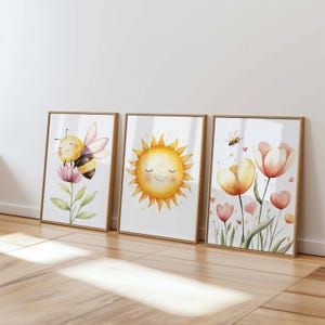 May include: Three framed watercolor art prints featuring a bee, a smiling sun, and flowers. The bee is yellow and black, the sun is yellow, and the flowers are pink and yellow. The frames are a light brown color.