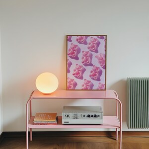 Pode incluir: Uma mesa lateral de metal rosa com uma l&acirc;mpada redonda e brilhante e uma impress&atilde;o emoldurada com um padr&atilde;o de ursinhos de goma rosa. Um sistema est&eacute;reo e livros est&atilde;o na prateleira inferior. A parede &eacute; branca e um radiador verde &eacute; vis&iacute;vel.