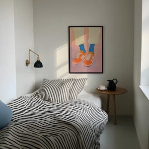 May include: A black and white striped bedspread with a black and white striped pillow on a bed in a white room. A framed painting of a pair of orange sneakers with blue socks hangs on the wall above the bed.