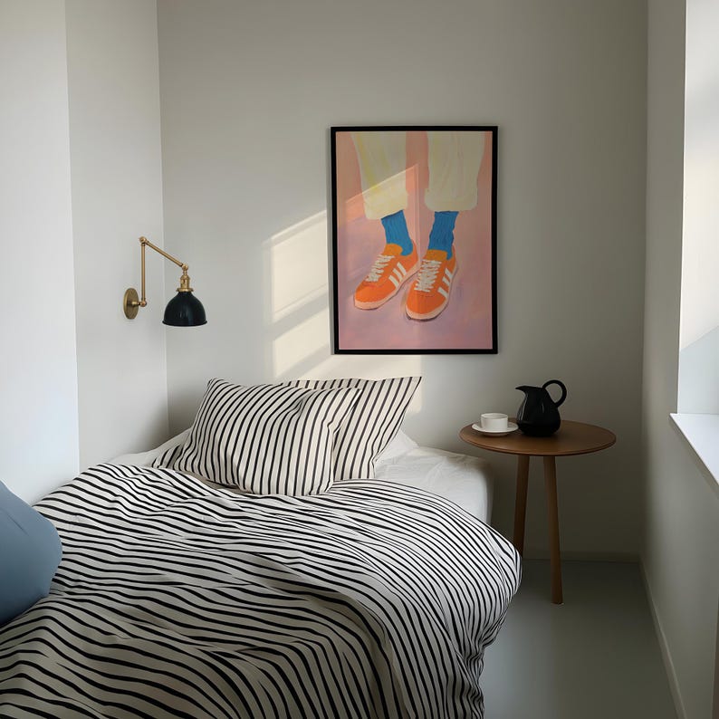 May include: A black and white striped bedspread with a black and white striped pillow on a bed in a white room. A framed painting of a pair of orange sneakers with blue socks hangs on the wall above the bed.