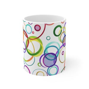 Può includere: Una tazza in ceramica bianca con un motivo ripetuto di cerchi sovrapposti in vari colori, tra cui rosso, verde, blu, viola, arancione e giallo. Il design copre la superficie esterna della tazza, che presenta un interno bianco semplice.