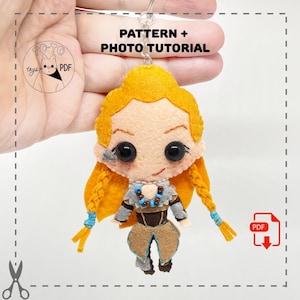 May include: A felt doll keychain featuring a character with yellow hair in braids, wearing a brown and gray outfit. The doll has large black eyes and a small smile. The keychain includes the text "PATTERN + PHOTO TUTORIAL".
