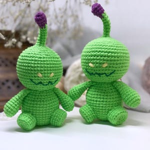 May include: Two green crocheted plush toys with purple tops and yellow eyes. The toys have a mischievous grin and are sitting on a white surface.