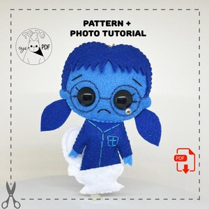 May include: A handmade blue felt doll with a sad expression, wearing glasses and a blue outfit. The doll has a white, cloud-like base. The image includes the text "PATTERN + PHOTO TUTORIAL" and a logo with a cat.