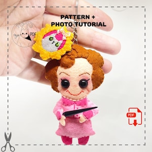 Puede incluir: Un llavero de fieltro hecho a mano con un personaje de dibujos animados con un vestido rosa, sosteniendo un puntero negro. El llavero incluye un dije en forma de flor amarilla con un diseño de gato. El texto en la imagen dice "PATTERN + PHOTO TUTORIAL".