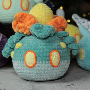 May include: Four crocheted plush toys in the shape of Pokemon. The toys are purple, teal, orange, and light blue. The teal toy has a yellow flower on top.