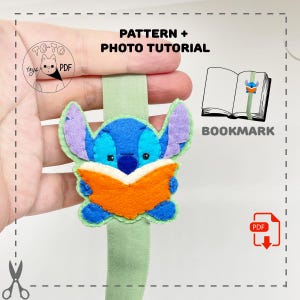 May include: A handmade felt bookmark featuring a blue cartoon character reading an orange book. The bookmark has purple ears and is attached to a green ribbon. The image includes the text "PATTERN + PHOTO TUTORIAL" and "BOOKMARK".
