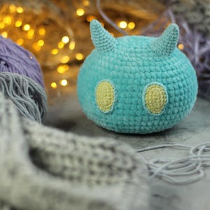 Puede incluir: Un juguete de peluche de color azul turquesa tejido a crochet con ojos amarillos y cuernos. El juguete está sentado sobre una superficie gris con hilo.