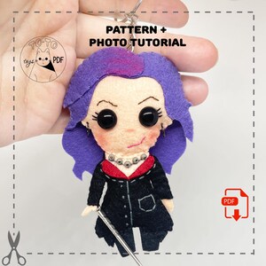 May include: A handmade felt doll keychain featuring a character with purple hair, large black eyes, and a black coat. The keychain includes the text "PATTERN + PHOTO TUTORIAL" and a PDF icon.