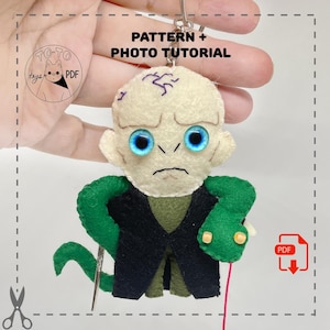 May include: A handmade felt keychain featuring a character with a pale face, blue eyes, and a green snake. The character wears a black coat and green vest. The text "PATTERN + PHOTO TUTORIAL" is visible.