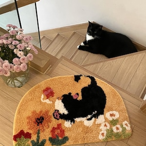 May include: A yellow half-circle doormat featuring a black and white cat design and colourful flowers. The cat is shown playing with a butterfly.