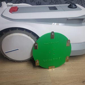 Puede incluir: Una pieza de repuesto de cuchilla circular verde para cortacésped robot con soportes de metal dorado. El texto "Made in Britain" y "EcoVacs GoCut G1" está impreso en la cuchilla.