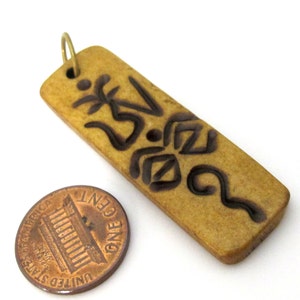 May include: A rectangular pendant with a carved design and a small gold-colored loop at the top. The pendant is a light brown color with dark brown carved symbols. A penny is next to the pendant for size comparison.
