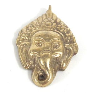 Puede incluir: Un colgante de lat&oacute;n detallado con la cara de Ganesha, el dios hind&uacute; de la sabidur&iacute;a. El colgante tiene un lazo en la parte superior para colgar y est&aacute; sobre un fondo blanco. El colgante mide aproximadamente 5 cm de altura.