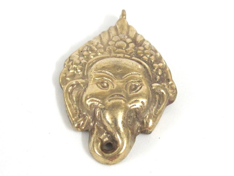 Puede incluir: Un colgante de lat&oacute;n detallado con la cara de Ganesha, el dios hind&uacute; de la sabidur&iacute;a. El colgante tiene un lazo en la parte superior para colgar y est&aacute; sobre un fondo blanco. El colgante mide aproximadamente 5 cm de altura.