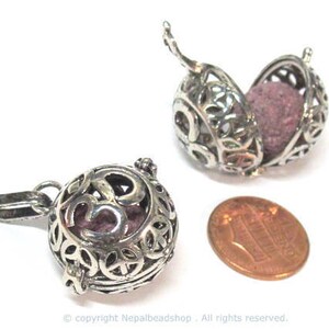 May include: Two silver-colored aromatherapy pendant necklaces with intricate openwork designs. One is open, revealing a purple insert. A penny is next to the pendants for size comparison. The pendants have a loop for a chain.