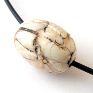 May include: An oval-shaped bead with a cream-colored base and dark brown veining, featuring carved vertical grooves along its surface. The bead is threaded onto a thin, smooth black cord that passes through its center horizontally.