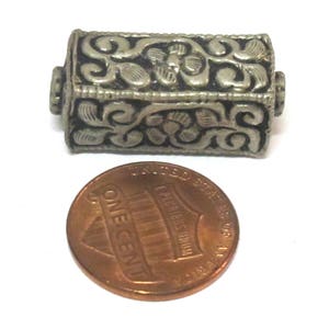 May include: A silver-colored, rectangular bead with intricate floral and scrollwork designs. The bead is shown next to a copper-colored one-cent coin for size comparison. The bead's detailed carvings are visible.
