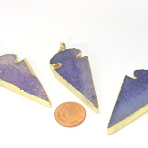May include: Three arrowhead-shaped pendants with a gold-colored trim and a purple, textured center. The pendants are displayed on a white background with a penny for size comparison. Each pendant has a small loop at the top.