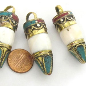 Puede incluir: Tres colgantes cilíndricos blancos, acentuados con detalles de latón, turquesa y rojo. Cada colgante tiene un lazo de latón en la parte superior para colgar. Los colgantes se muestran sobre una superficie blanca.