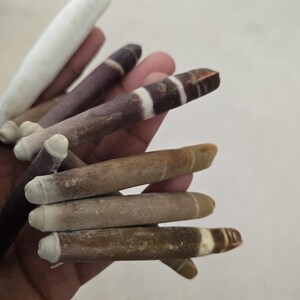 May include: A collection of handmade, cylindrical objects in varying shades of brown, tan, and white. Some have a white tip and a striped pattern. The objects are held in a hand against a neutral background.