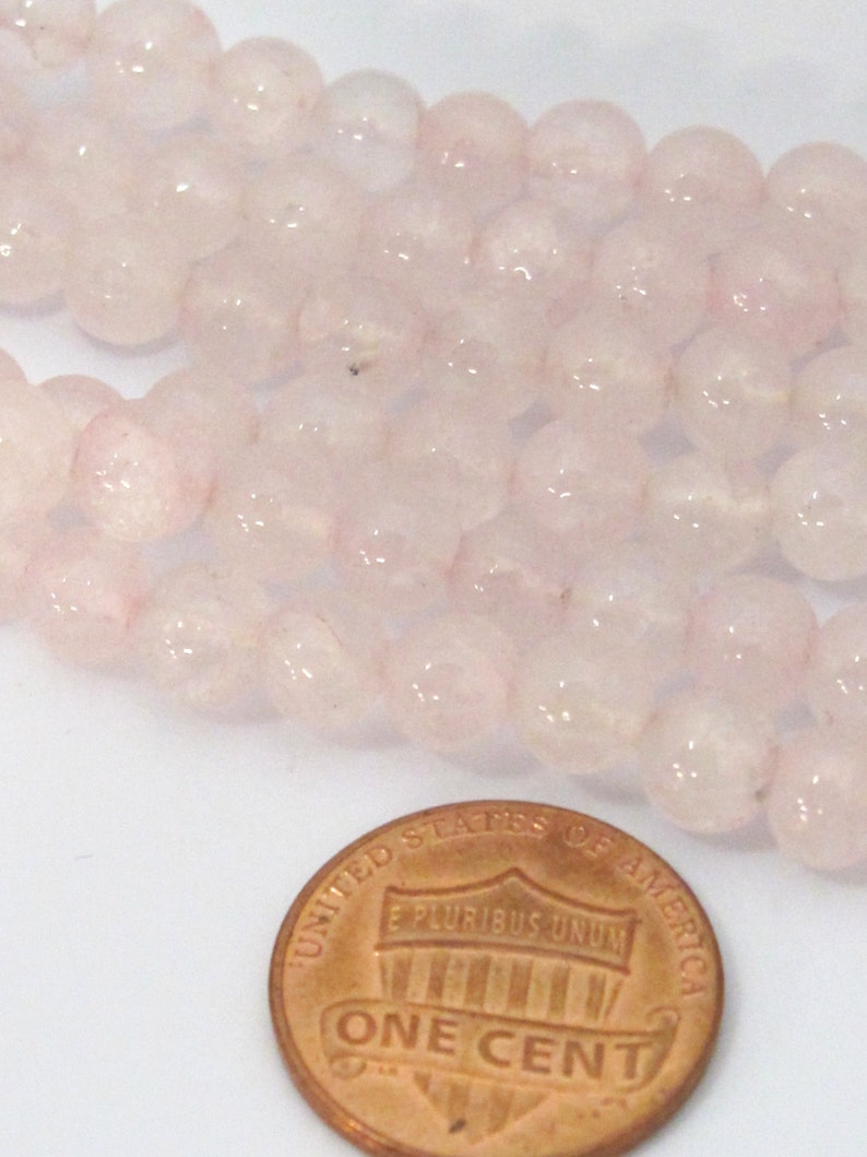 Pu&ograve; includere: Primo piano di perline di quarzo rosa rosa chiaro. Le perline sono rotonde e traslucide. Una moneta di rame da un centesimo &egrave; in primo piano per il confronto delle dimensioni.