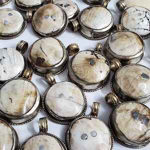 May include: A collection of round pendants with a cream-colored, textured surface and silver-colored metal rims. Each pendant has a small loop for a chain. The pendants are arranged closely together, showcasing their unique patterns and textures.