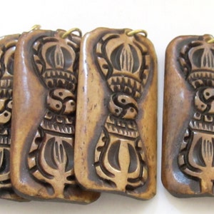 May include: Four rectangular pendants with a carved design. The pendants are a light brown color with a darker brown carved design. Each pendant has a gold-colored loop at the top.