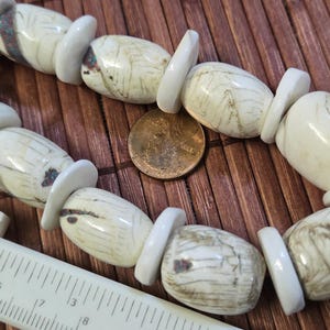 May include: A strand of off-white and gray stone beads with white spacers. A penny and a white ruler are next to the beads on a brown bamboo mat. A black cord is visible at the top.