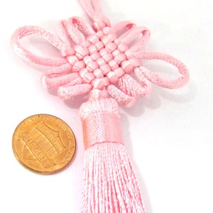 May include: A pink Chinese knot, also known as a decorative knot, with a tassel. The knot is made of woven pink satin cord and has a symmetrical design. A US one-cent coin is next to the knot for size comparison.