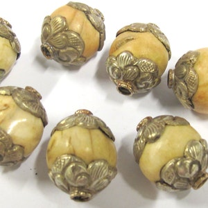May include: Close-up of seven ivory-colored, oval-shaped beads with ornate, antique-style metal caps. The caps feature a floral design and are a tarnished gold color. The beads have a smooth, polished surface.