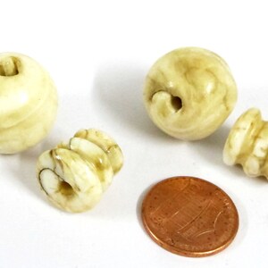 May include: A collection of off-white beads with brown marbling. The beads are various shapes, including round and cylindrical, with holes for stringing. A penny is included for size reference.