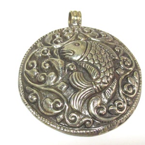 May include: Silver-toned pendant with a detailed relief of a fish within a circular frame. The fish is surrounded by swirling, ornate patterns. The pendant has a loop at the top for a chain.