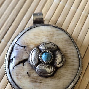 May include: A round pendant with a silver frame and a central flower design. The flower has a turquoise stone in the center. The pendant is set on a light-colored, textured background.