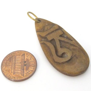 May include: A brown, teardrop-shaped pendant with a carved Om symbol and a small brass loop. The pendant is next to a United States one-cent coin.