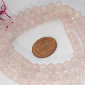 Pu&ograve; includere: Fili di perline di quarzo rosa pallido. Le perline sono rotonde e lucide. Una moneta &egrave; accanto alle perline per il confronto delle dimensioni. Una nappa rosa &egrave; attaccata all'estremit&agrave; dei fili.