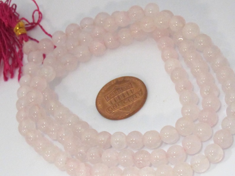 Pu&ograve; includere: Fili di perline di quarzo rosa pallido. Le perline sono rotonde e lucide. Una moneta &egrave; accanto alle perline per il confronto delle dimensioni. Una nappa rosa &egrave; attaccata all'estremit&agrave; dei fili.