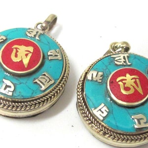 May include: Two round turquoise pendants with gold-colored metal accents. Each pendant features a central red circle with a gold symbol and is surrounded by silver-colored symbols. The pendants have a braided metal edge.
