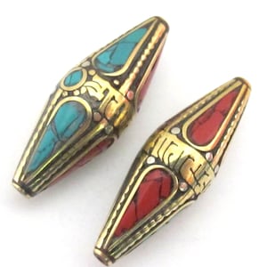 May include: Two decorative beads with a gold-colored metal casing. One bead features turquoise and red stone inlays, while the other has red stone inlays. The beads have an elongated, symmetrical shape with intricate metalwork.