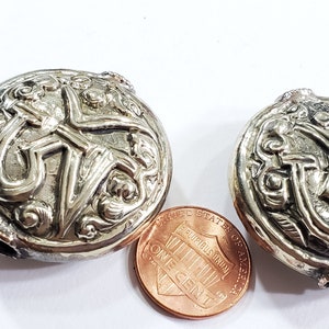 May include: Two silver-colored, round, ornate buttons with detailed relief patterns. A copper penny is placed next to the buttons for size comparison. The buttons have a vintage or antique aesthetic.