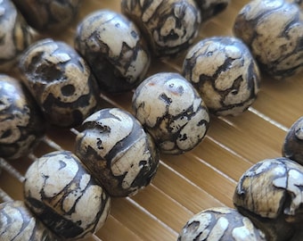 Può includere: Primo piano di una fila di grani di preghiera. Le perline sono di colore beige chiaro con motivi vorticosi marrone scuro. Le perline sono disposte in file su una superficie strutturata marrone chiaro.