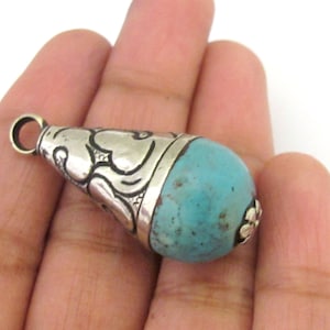 May include: A silver-toned pendant with a turquoise stone. The pendant has a teardrop shape, with intricate etched designs on the silver cap and a small loop for a chain. The turquoise stone is a vibrant blue-green color.