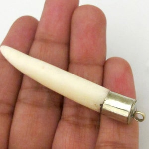 May include: Ivory-colored pendant with a tapered shape and a silver-colored metal cap. The pendant has a small loop at the end of the cap. The pendant is held in a hand.