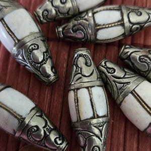May include: Close-up of several decorative beads with a white center and ornate silver-colored metal ends. The beads are arranged on a reddish-brown surface, with a penny visible in the lower left corner.