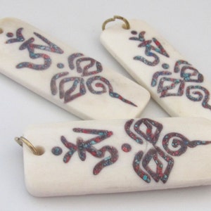 May include: Ivory-colored rectangular pendants with a brass loop. Each pendant features a design in red and turquoise, including the Om symbol and floral patterns. The pendants are likely for jewelry or craft projects.