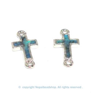 May include: Two silver-toned cross charms with a teal and iridescent inlay. Each cross has a loop at the top and bottom for attaching to jewelry. The crosses are set against a white background.